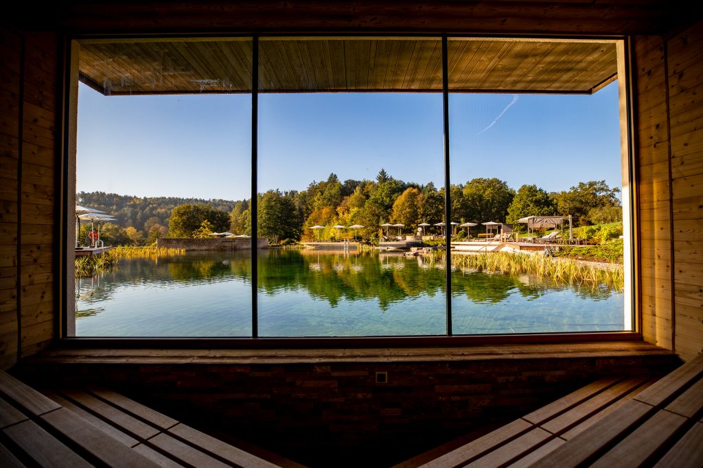 Direkt am türkisblauen, 70 Meter langen Naturbadeteich „kleiner Wasgausee“ im Wald- und Wiesenareal des Pfalzblick Wald Spa Resort, genießen Erholungssuchende in der finnischen Sauna den Panoramablick 
